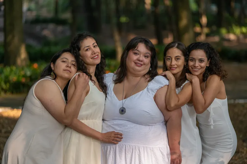 Group portrait of five women in white dresses beneath trees in warm light